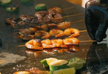 Close-up of a chef cooking seasoned shrimp skewers on a sizzling hot flat-top grill. 